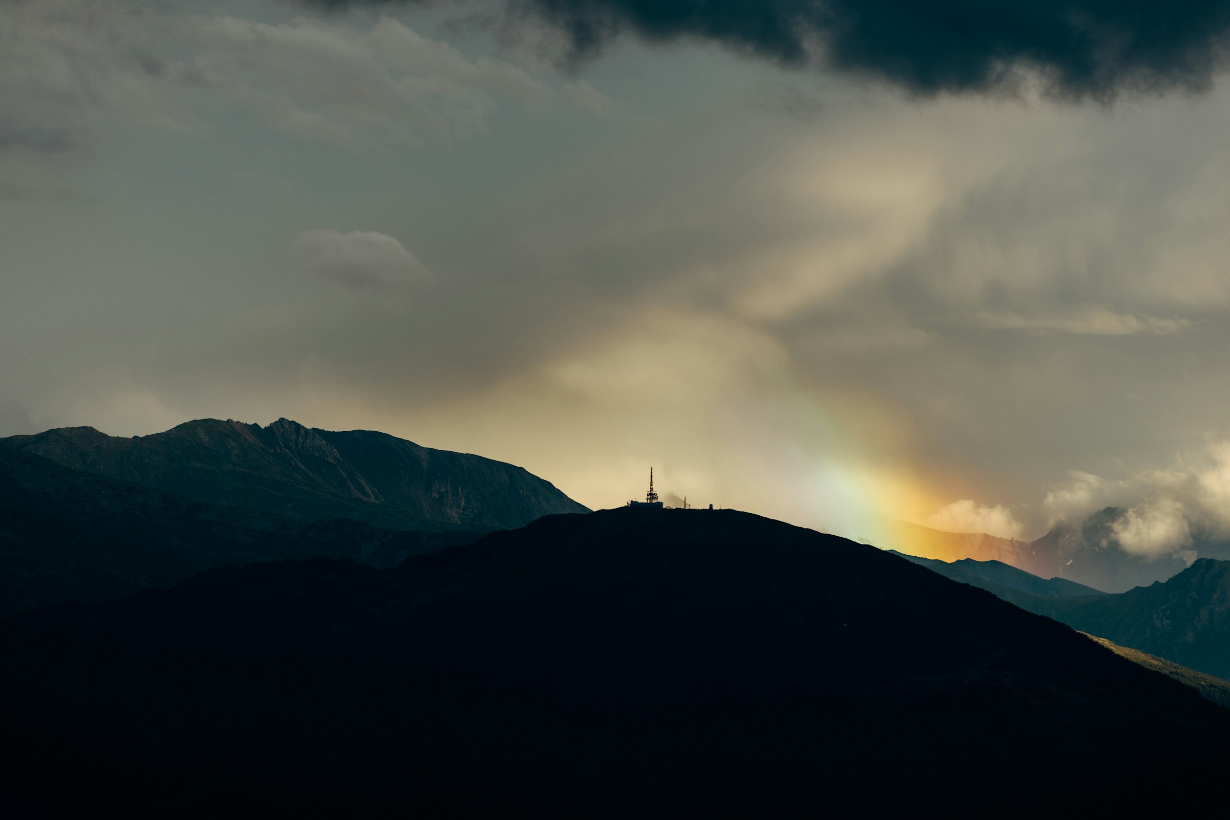 A dark mountain silhouette with a faint rainbow and a communication tower by Florian Schindler