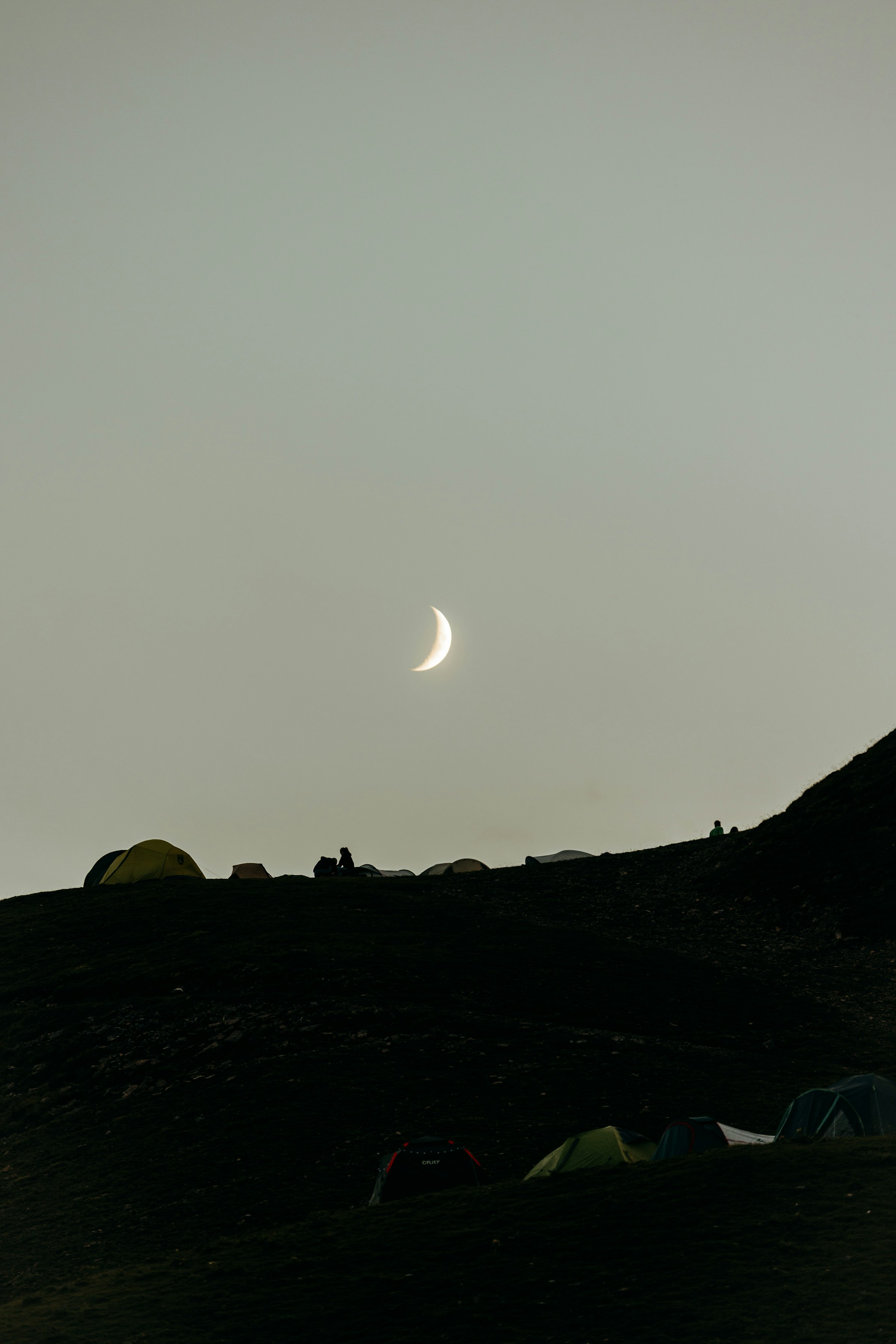 A crescent moon over dark hills with tents by Florian Schindler