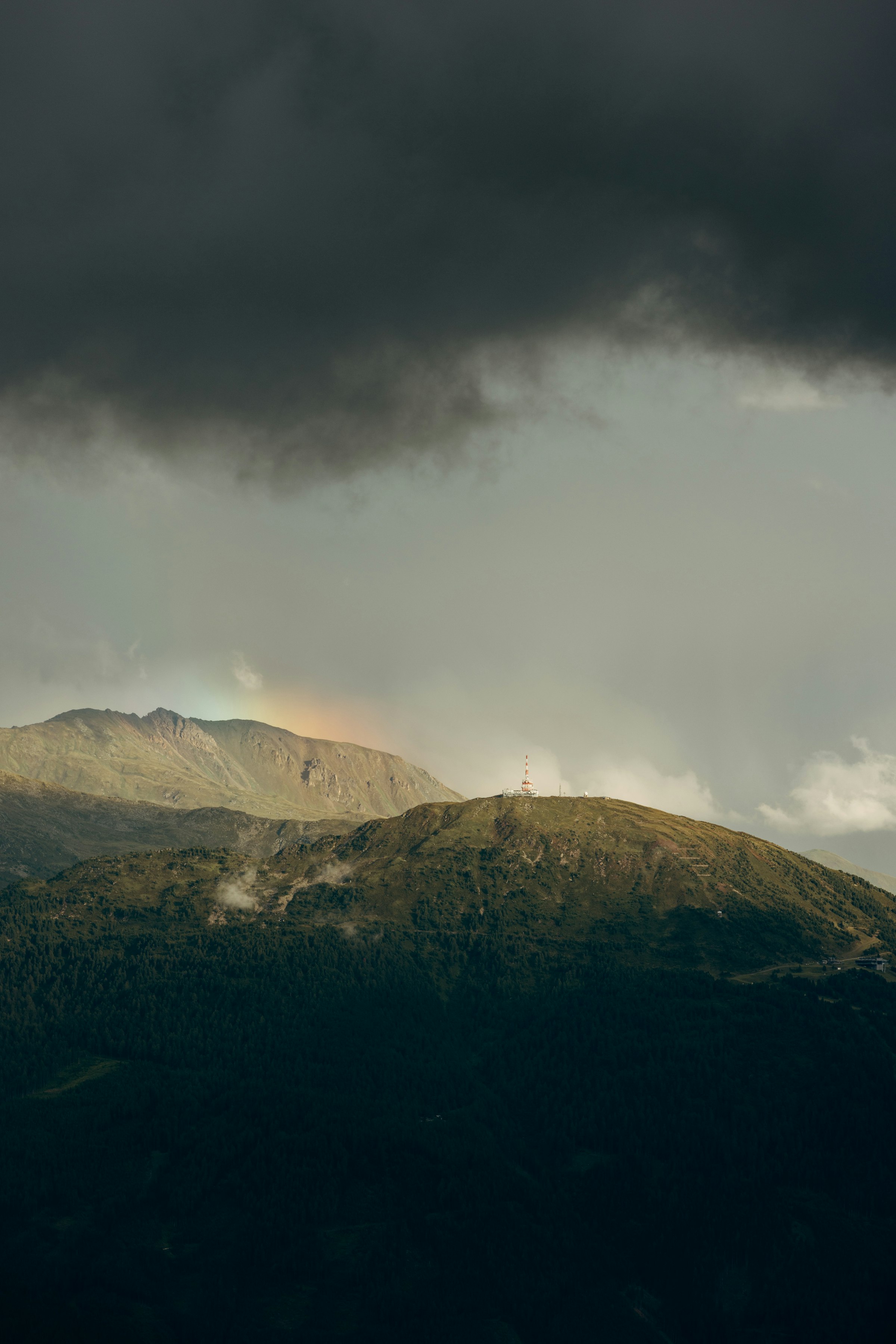 Dark clouds above sunlit mountains with a subtle rainbow by Florian Schindler