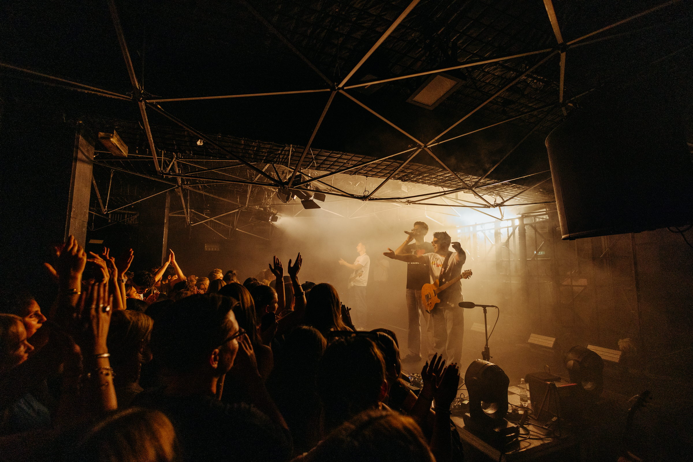 A band on stage in warm amber haze while the crowd raises their hands by Florian Schindler