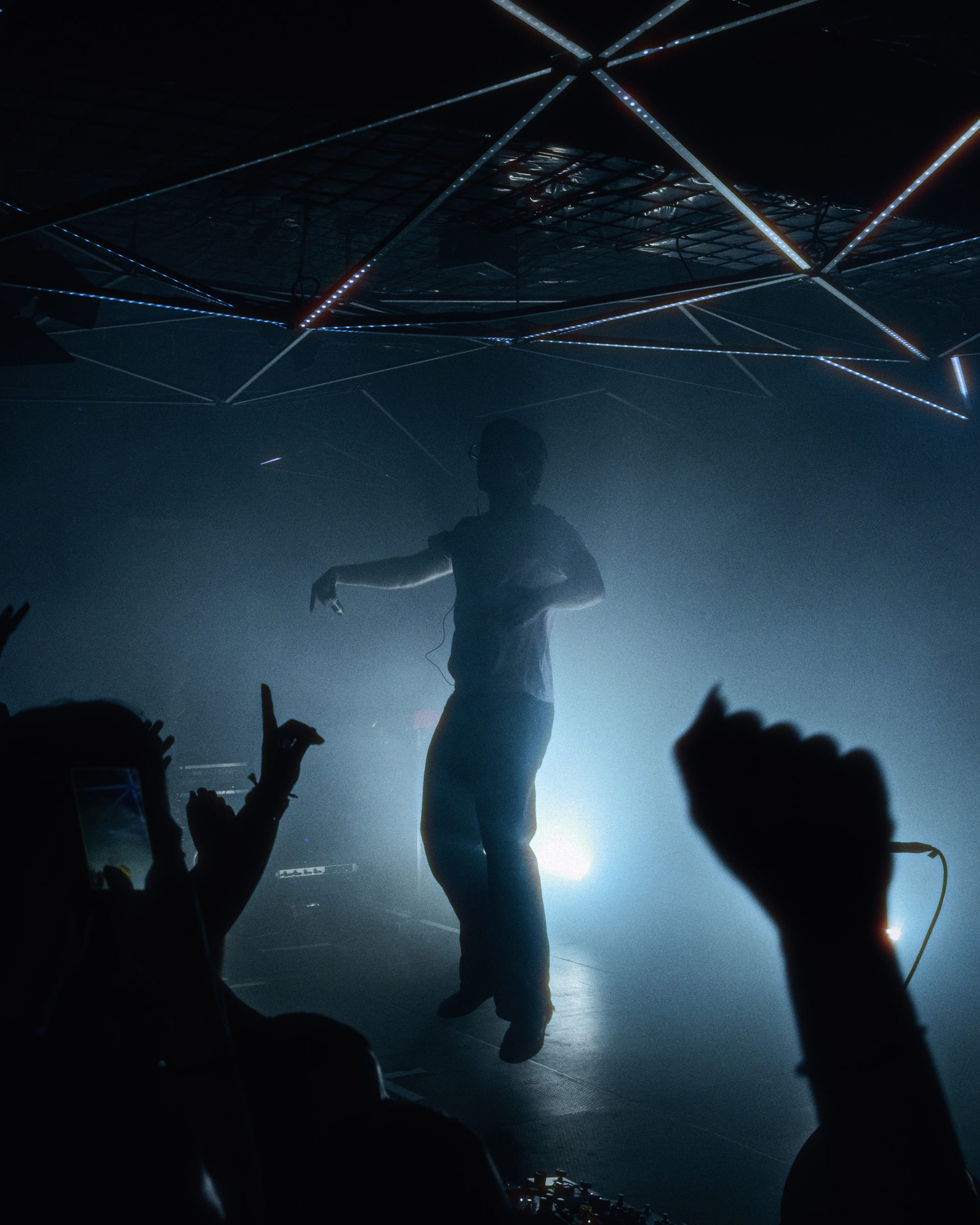A performer silhouetted in cold backlight and fog with fans raising their hands in the foreground by Florian Schindler